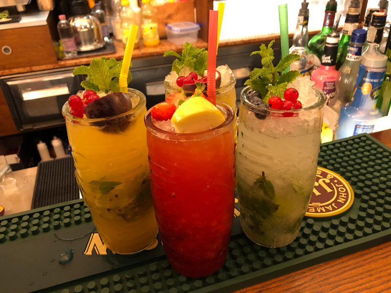 Tre cocktail colorati con guarnizioni di frutta fresca e menta, allineati su un bancone da bar in legno